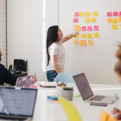 Woman delivering a training session for a small group. She places sticky notes on a large whiteboard, while the group sit at a table working on laptops.